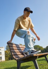 En person som balanserer på en benk i tre med en metallramme. De har på seg en beige skjorte, lyseblå slitte jeans og svarte joggesko.