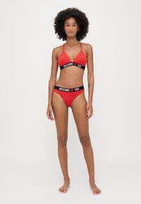 Costum de baie roșu format dintr-un top triunghiular și un slip cu talie joasă. Accesorizat cu benzi negre "MOSCHINO YOU". Textură netedă, design sport.