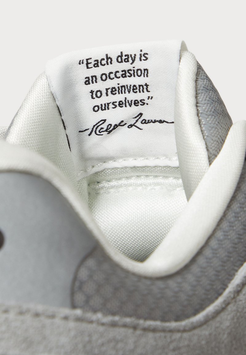 Graue Sneaker mit einer strukturierten Oberseite, glattem Innenfutter und einem genähten Zungenetikett mit einem bedruckten Zitat.