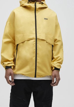 Yellow hooded jacket with black zipper and logo, worn with black cargo pants and white shirt underneath, shown from mid-torso down.