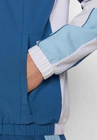 Treningsjakke i livlig blått og hvitt, med teksturert stoff, elastiske ermer og en skjult sidelomme med glidelås.