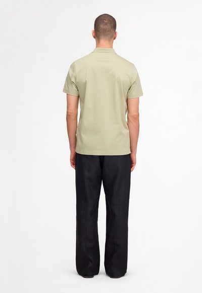 Man staand met zijn rug naar de camera, draagt een beige kortarmig overhemd en losse zwarte broek tegen een effen witte achtergrond.