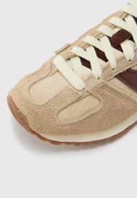 Béžová semišová teniska s textúrovaným povrchom, krémovými šnúrkami a hnedými koženými doplnkami. Zaoblená špička a gumová podrážka s jemným leskom.