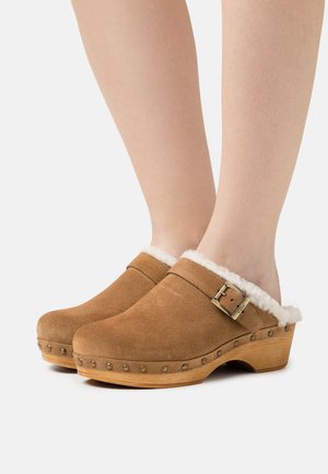 Brown suede clogs with a faux fur collar, wooden platform, and decorative metal studs along the edge. Closed-toe design with buckle detail.