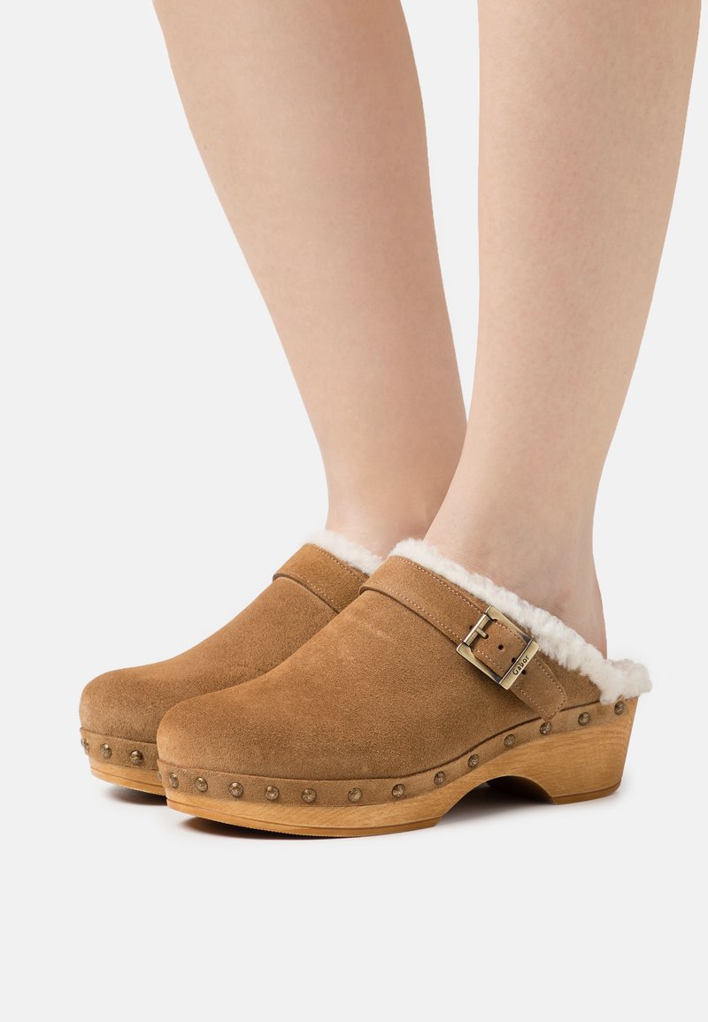 Braune Wildleder-Clogs mit einem Kunstfellkragen, hölzernen Plattformen und dekorativen Metallnieten entlang des Randes. Geschlossene Zehenform mit Schnallen-Detail.