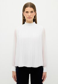 Weiße Bluse mit hohem Kragen, langen durchsichtigen Ärmeln und vertikalen Falten. Der Saum ist geschwungen, mit knöpfbaren Manschetten und einer glatten Textur.