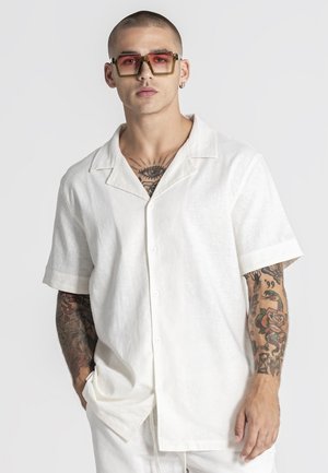 Mann mit Tätowierungen auf Armen und Brust, der große quadratische braune Sonnenbrillen und ein weißes kurzärmeliges Hemd mit Knopfleiste und Tasche trägt.