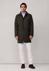 Oliivinvihreä, nappikiinnitteinen takki, jossa on räätälöity muotoilu, pitkät hihat ja suora leikkaus. Päällä vaaleansinisen paidan kanssa, yhdistetty valkoisiin housuihin.