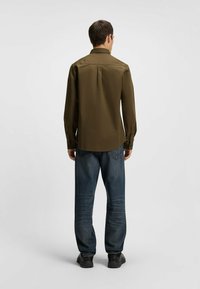 Olive green long-sleeve shirt, button-down collar, straight hem. Worn with dark blue jeans featuring a faded pattern and black shoes.