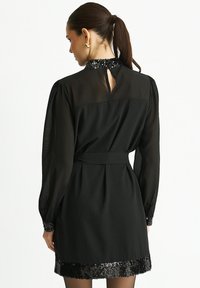 Black dress with long sleeves, semi-sheer texture, sequined collar and hem, belted waist, and back keyhole opening.