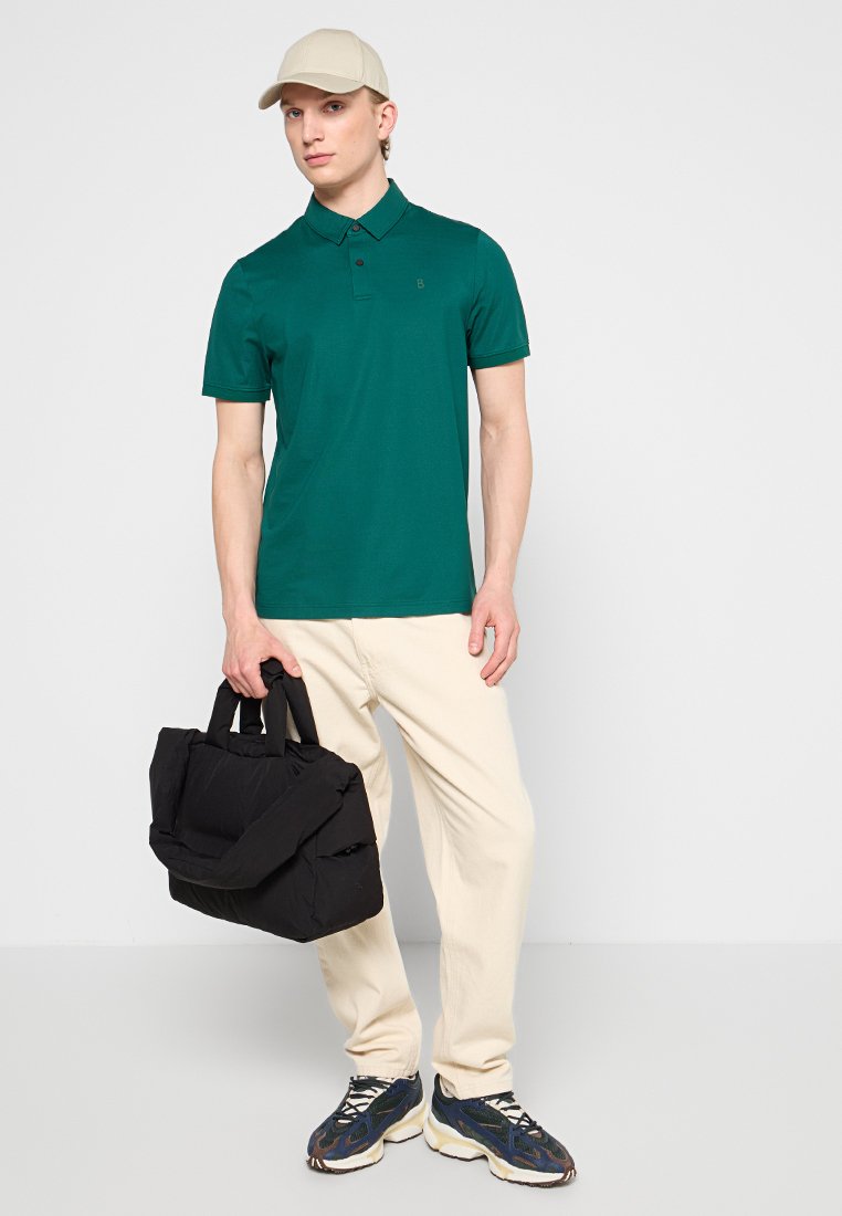 Mann trägt eine beigefarbene Mütze, ein grünes Polohemd, cremefarbene Hose und Turnschuhe, hält eine schwarze gepolsterte Tasche an den Griffen und steht vor einem weißen Hintergrund.