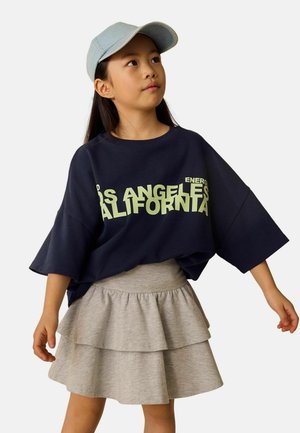 Ung pige iført en lyseblå kasket, mørkeblå oversized T-shirt med teksten "Los Angeles California" og et gråt lagdelt skørt, der kigger opad.