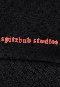 Zwarte stof tentoongesteld met de tekst "spitzbub studios" in vetgedrukte, rode letters. Het materiaal lijkt gebreid en heeft een gladde textuur.