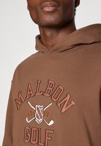 Brūns kapuce ar izšūtu "Malbon Golf" logo oranžā un krēmkrāsā, ar krustotiem golfa nūjām. Mīksta auduma, parasts piegriezums.