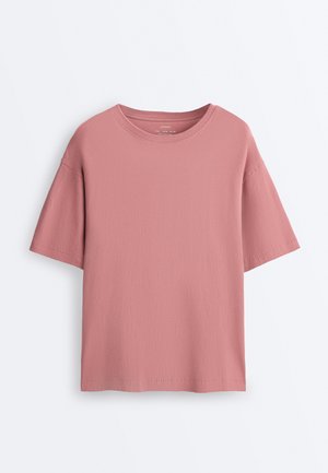 Prašno roza bombažna majica s kratkimi rokavi, okrolim izrezom in sproščenim krojem, prikazana na enobarvnem belem ozadju.