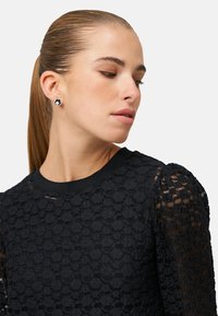 Fekete csipke felső virágmintával és felfújt ujjakkal. A modell fekete tüske fülbevalót visel. Sima haj alacsony lófarokban, semleges háttér.