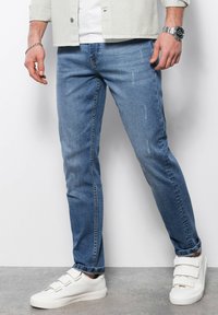 Vaqueros de denim azul claro con un corte ajustado, que presentan un ligero desgaste y arrugas. Combinado con zapatillas blancas con correas de velcro.