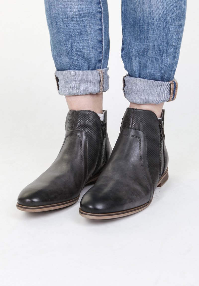Personne portant un jean bleu retroussé et des bottines en cuir noir avec fermetures éclair latérales sur fond blanc.
