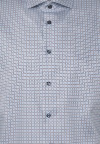 Gros plan d’une chemise boutonnée avec un motif géométrique aux tons bleus, blancs et bruns, avec des boutons noirs sur le devant.