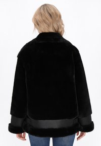Persona bionda che indossa una giacca in pelliccia ecologica nera con rifiniture in pelle e jeans blu, vista da dietro su uno sfondo bianco.