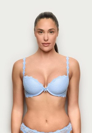 Donna con capelli scuri raccolti, che indossa un reggiseno di pizzo azzurro chiaro e slip coordinati, in piedi davanti a uno sfondo bianco uniforme.