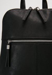 Sac à dos en cuir noir avec fermeture éclair argentée et double tirettes, avec une poche avant zippée munie d'un pompon en cuir.
