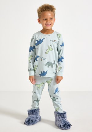 Junges Kind lächelt, trägt hellblaue Pyjamas mit Dinosaurier-Print und flauschige blaue Hausschuhe mit Krallen, steht vor weißem Hintergrund.