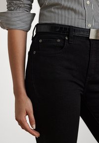 Jeans in denim nero con vestibilità slim, caratterizzati da tasche frontali e rivetti metallici. L'individuo indossa una cintura nera con texture e una camicia a righe.