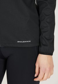 Schwarze Sportjacke mit einer glatten, matten Oberfläche. Mit dem "ENDURANCE"-Logo in Weiß am Saum. Langarm mit elastischen Bündchen.