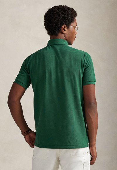 Zelena polo majica s kratkimi rokavi, enobarvna tekstura, ravnim ovratnikom in ravnim robom, nošena z belimi hlačami. Prikazan zadnji pogled.