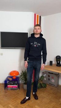 Granatowa bluza z kapturem z logo "Tommy HILFIGER", niebieskie dżinsy i ciemne loafersy. Na ramionach widoczne tatuaże. W tle kolorowa ściana i meble.