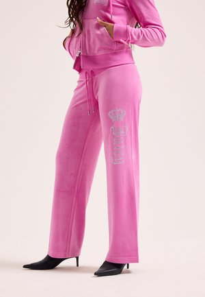 Donna che indossa pantaloni in velluto rosa acceso con la scritta "Juicy" e un disegno di corona sulla gamba, abbinati a una felpa con cappuccio coordinata con zip e tacchi neri a punta.