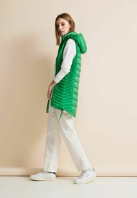 Zelená, nadýchaná bezrukávová vesta s kapucí a vzorem kosočtverců, spárovaná s bílým dlouhým rukávem a světlými kalhotami.