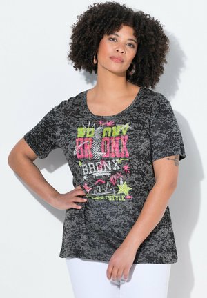 Kobieta o kręconych włosach, ubrana w czarną koszulkę z kolorowym napisem Bronx i białe spodnie, pozująca na białym tle.