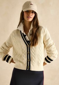 Kreemitooniline nööbiga kardigan, millel on V-kaelus ja tumesinised triibud, nelja suure nööbi ja narmastega mansettidega, koos kollase särgi ja mütsiga.