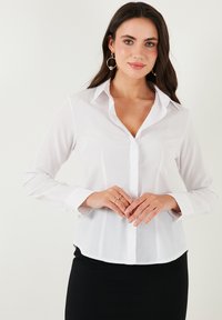 White long-sleeve button-up shirt with a pointed collar, front placket, and curved hem. Made from smooth, lightweight fabric.