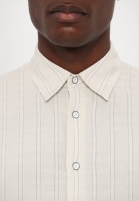 Light-colored shirt with vertical blue stripes, button-up collar, and rounded edges. Features white buttons with a slight sheen.