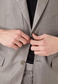 Blazer gris claro con textura sutil, que presenta un cierre de dos botones. Manos ajustan el botón de la solapa, revelando una camiseta negra en el interior.