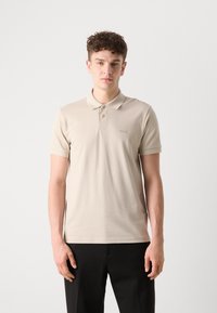 Beige polo skjorte med krage, tre knapper foran og brodert logo. Korte ermer og myk, glatt tekstur. Kombinert med svarte bukser.