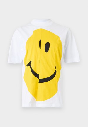Chemise blanche à manches courtes avec un grand visage souriant jaune imprimé sur le devant, montrant des yeux noirs et un sourire courbé.