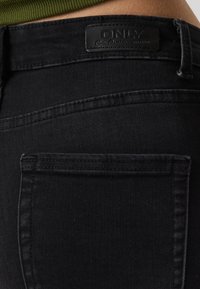 Jeans de mezclilla negra con una textura suave, que presentan un bolsillo trasero y una etiqueta de marca rectangular de cuero negro en relieve.