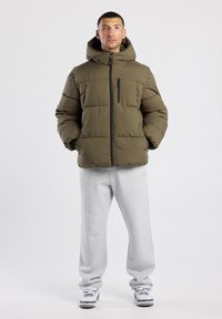 Olive green padded jacket with a hood, featuring a front zipper and a chest pocket. Worn with gray sweatpants and white sneakers.