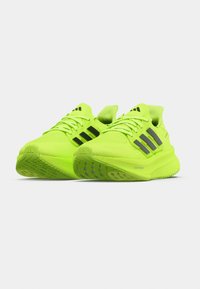 Een paar fel neon groene Adidas hardloopschoenen met zwarte strepen en gestructureerde zolen, naast elkaar geplaatst op een witte achtergrond.