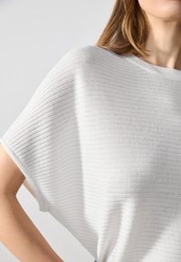 Biały, prążkowany sweter o luźnym kroju, z szerokimi rękawami i gładką teksturą. Prosty design, minimalne zdobienia i miękkie wykończenie.