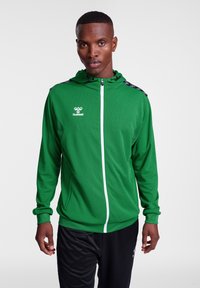 Grøn zip-up hoodie lavet af letvægtsmateriale, med en hvid lynlås, sorte detaljer på skuldrene og et logo på brystet.