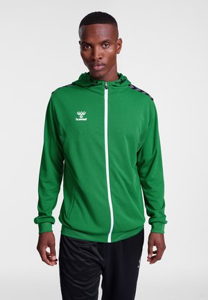 Veste à capuche zippée verte en matériau léger, avec une fermeture éclair blanche, des accents noirs sur les épaules et un logo sur la poitrine.