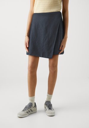 Nainen, jolla on tumma sininen kietaisutyylinen minihame, kerma valkoinen hihaton toppi, valkoiset sukat ja harmaat Adidas-tossut, seisoo valkoisella taustalla.