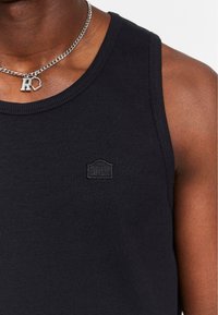 Black ribbed tank top featuring a round neckline, thick shoulder straps, and a small black logo patch on the chest. Silver chain necklace with pendant.
