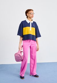 Navy and yellow color-blocked polo shirt with a white collar, paired with pink wide-legged trousers. Model holds a pink backpack.
