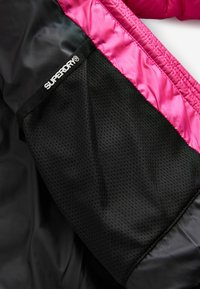 Čierna a ružová bunda s lesklým povrchom; má čierny sieťový rukáv; na manžete je zobrazené logo "SUPERDRY®".